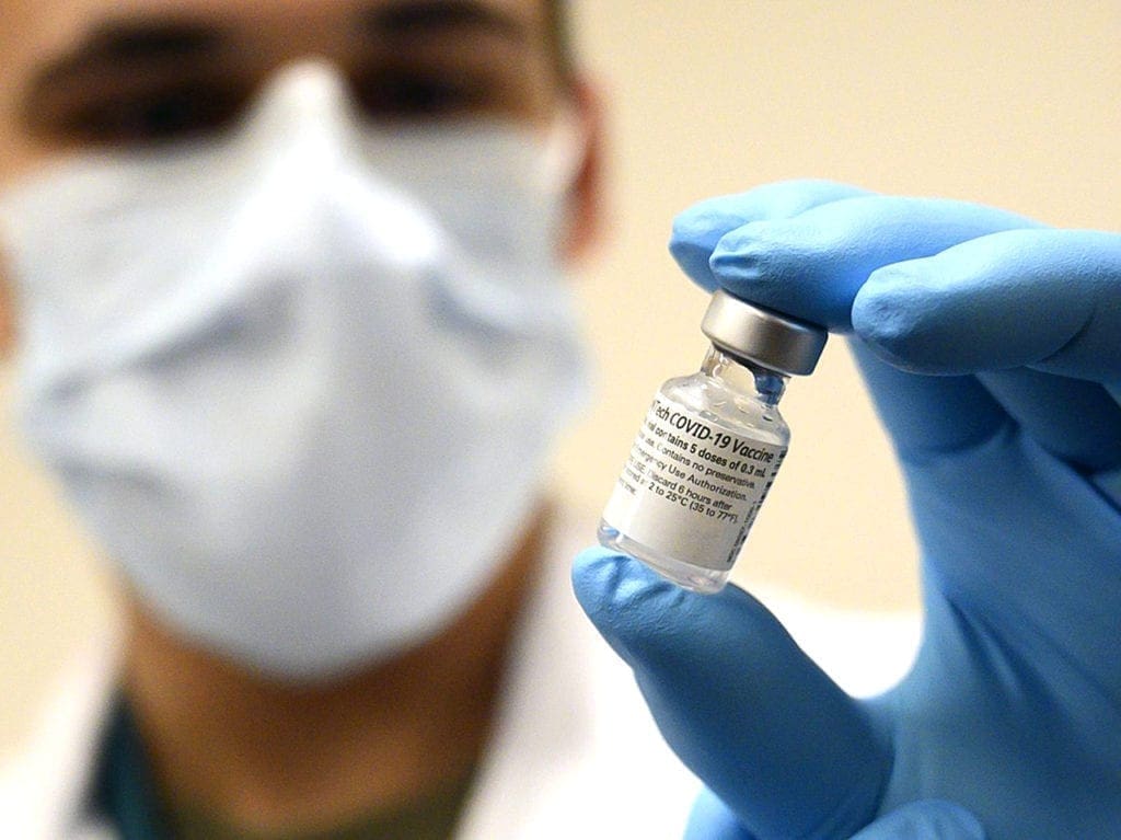 A man in a mask with a blue surgical glove holds a vial of a COVID-19 vaccine.