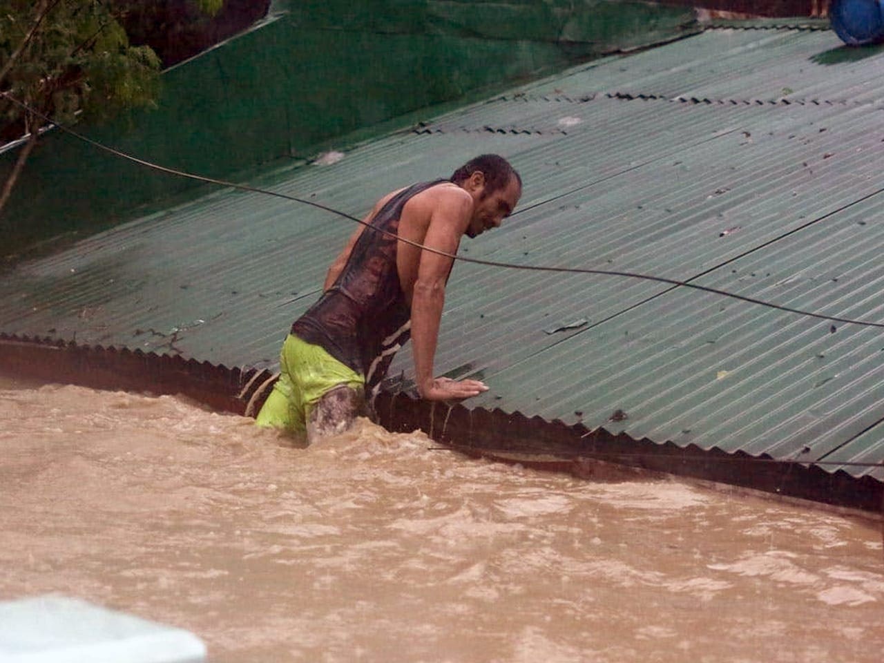 A man lifts himself onto a roof as his legs are submerged in flood water.